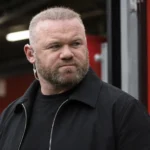 Wayne Rooney kijkt ernstig in een overdekte voetbalstadiongang met een Manchester United-plaque op de achtergrond.