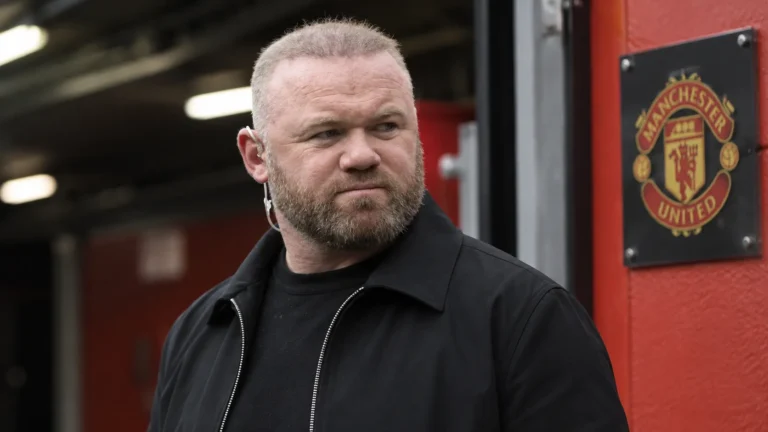 Wayne Rooney kijkt ernstig in een overdekte voetbalstadiongang met een Manchester United-plaque op de achtergrond.