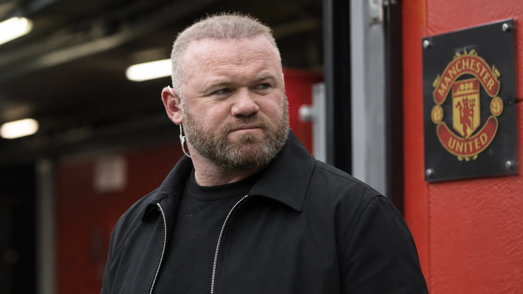 Wayne Rooney kijkt ernstig in een overdekte voetbalstadiongang met een Manchester United-plaque op de achtergrond.