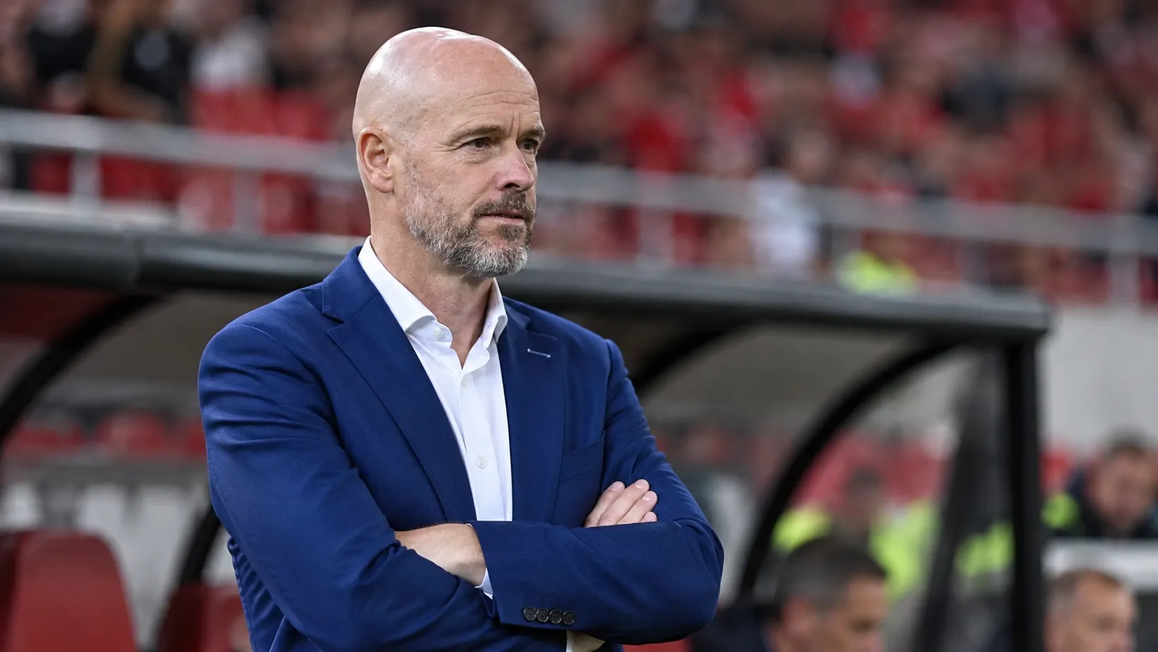 Erik ten Hag in een blauw pak aan de rand van een voetbalveld.