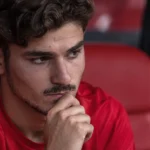 Een voetballer in een rood shirt zit geconcentreerd op de bank naast het veld.