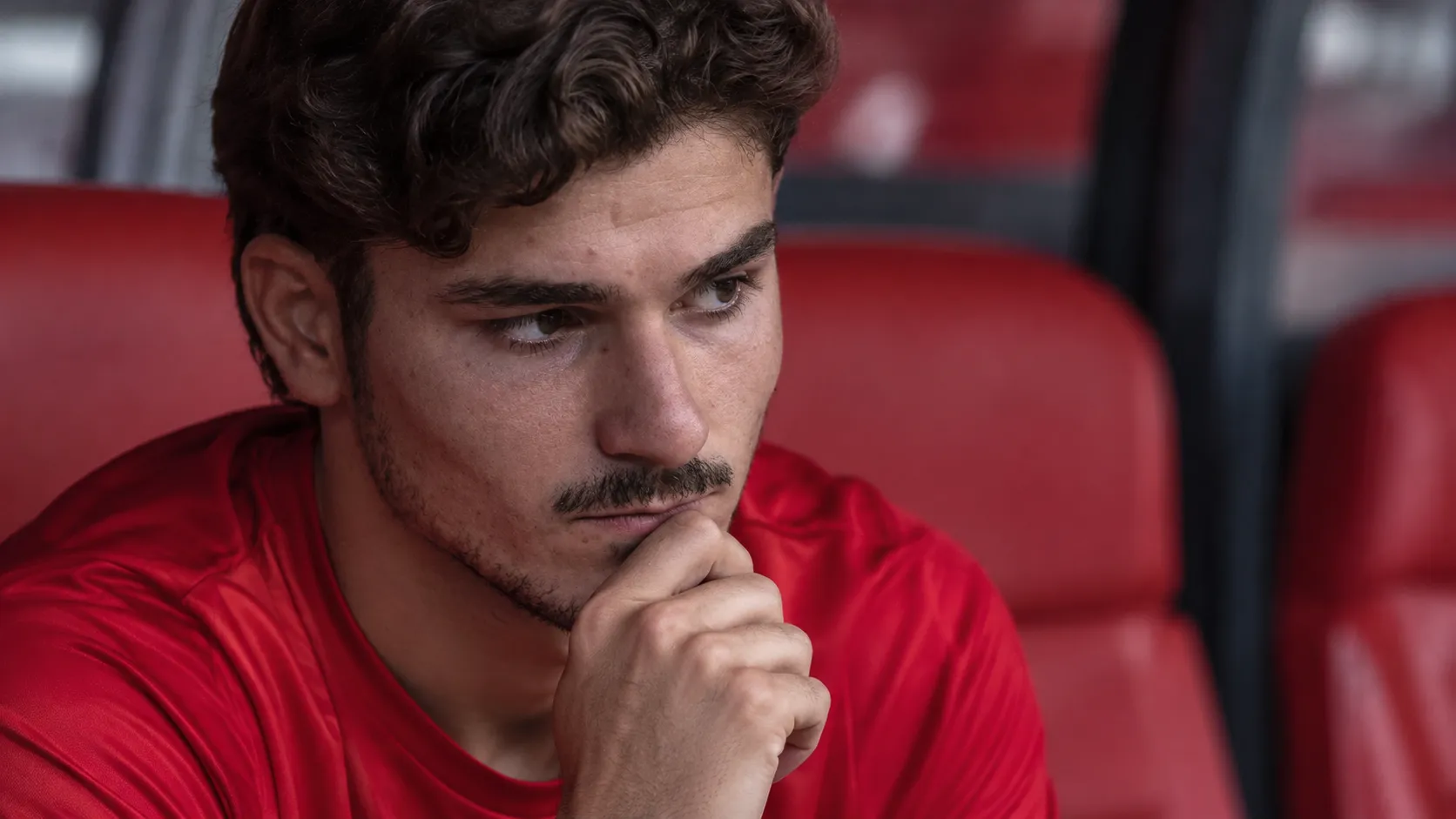 Een voetballer in een rood shirt zit geconcentreerd op de bank naast het veld.