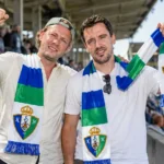 Twee Sporting Hasselt-supporters met blauw-groen-witte clubssjaals op de tribune.