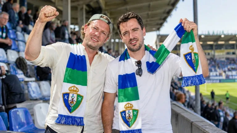 Twee Sporting Hasselt-supporters met blauw-groen-witte clubssjaals op de tribune.