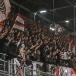 Een groep RC Lens-supporters achter een veiligheidsnet in een avondstadion.