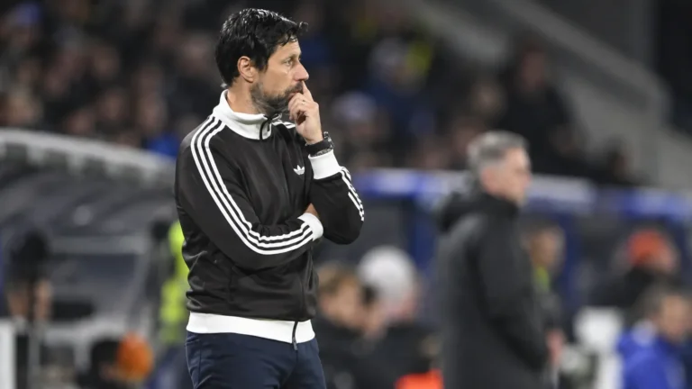 Een coach in een zwarte trainingsjas langs de zijlijn van een voetbalveld.