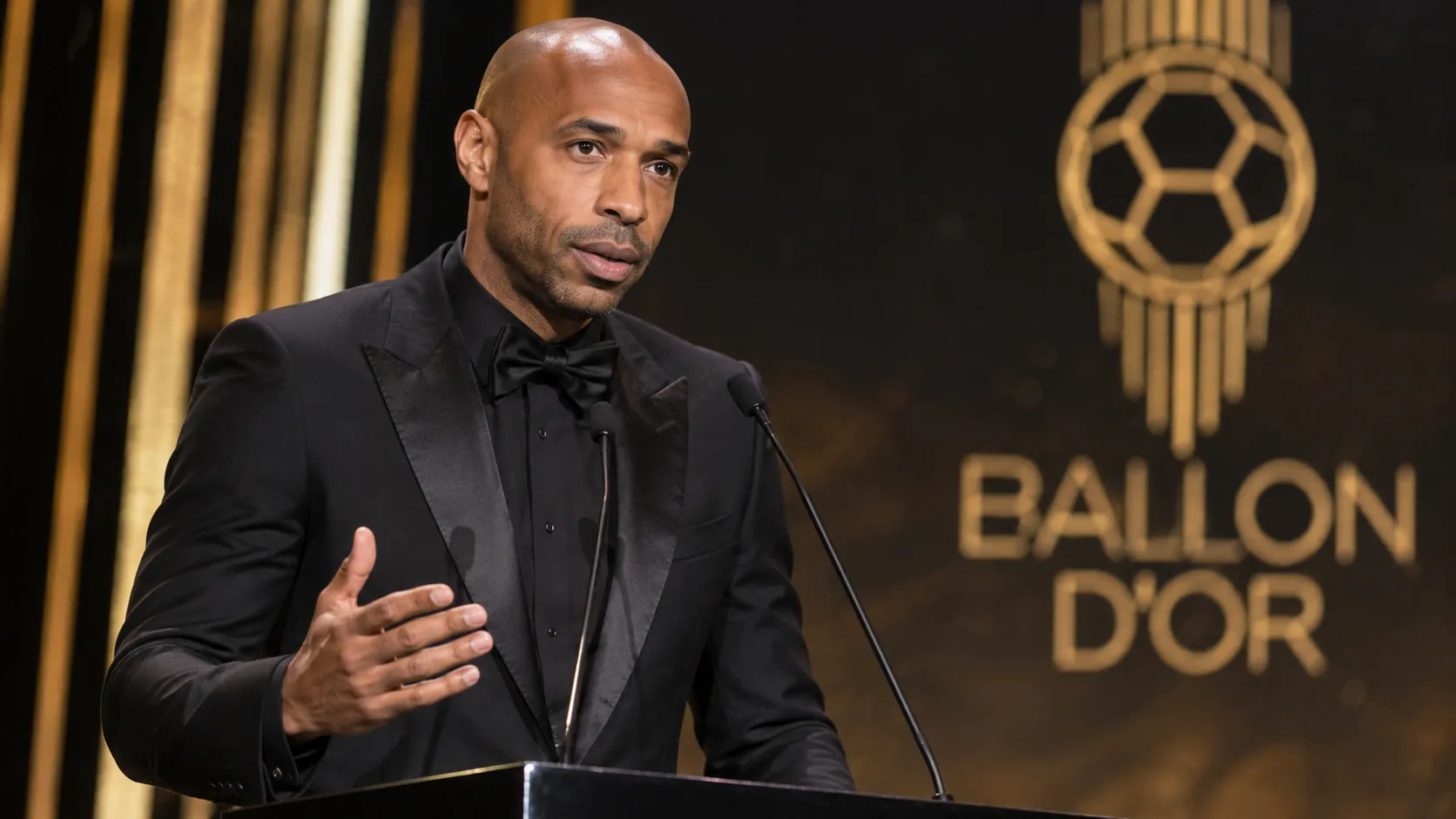 Thierry Henry spreekt op een Ballon d'Or-podium met een subtiel evenementsscherm op de achtergrond.