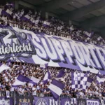 Een volle groep Anderlecht-supporters met paars-witte vlaggen en een groot spandoek in de tribune.