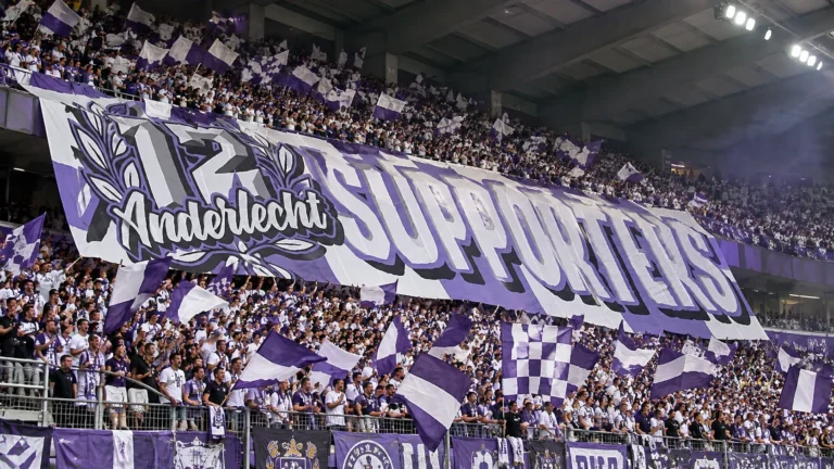 Een volle groep Anderlecht-supporters met paars-witte vlaggen en een groot spandoek in de tribune.