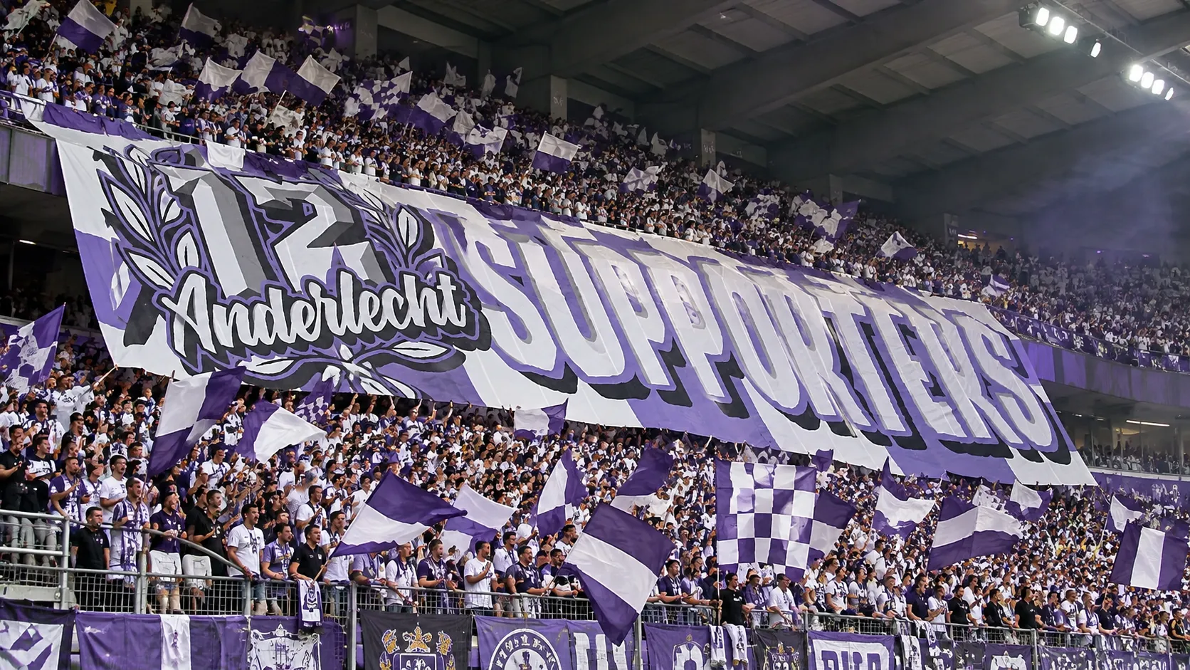 Een volle groep Anderlecht-supporters met paars-witte vlaggen en een groot spandoek in de tribune.