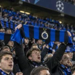 Collectieve Club Brugge-supporters met blauw-zwarte sjaals in een verlicht stadion.