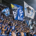 KRC Genk-supporters in blauwe kledij zwaaien met grote vlaggen op de tribune.