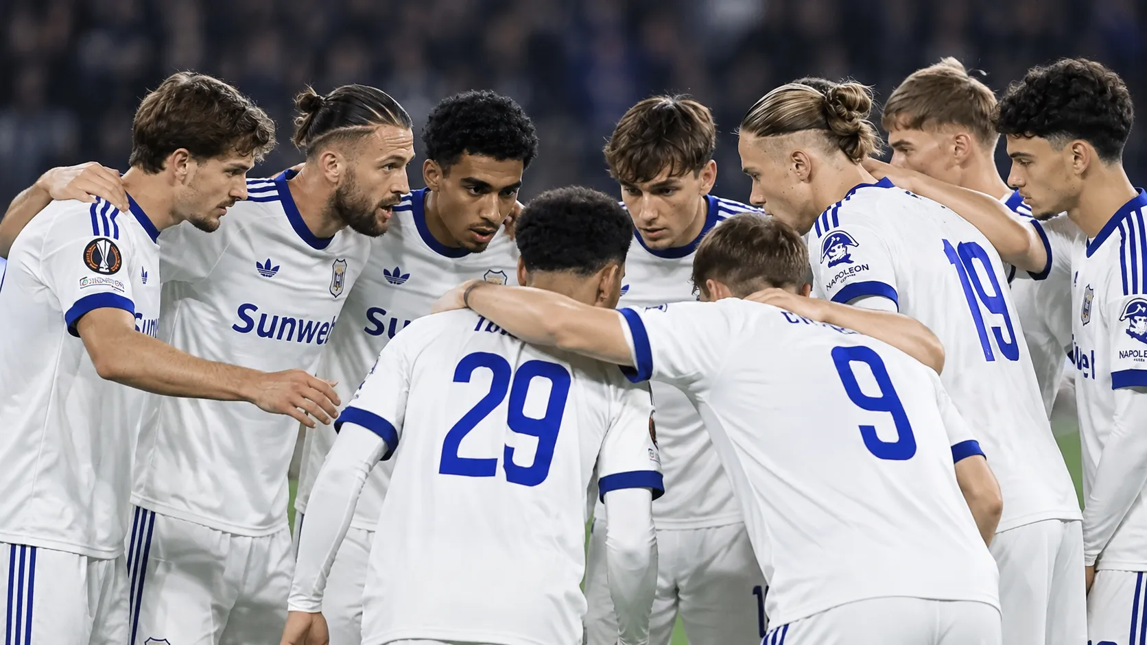 Een groep voetballers in witte tenues vormt een hechte huddle op het veld.