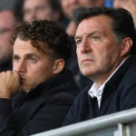 Twee niet-spelende clubvertegenwoordigers zitten aandachtig op de tribune van een voetbalstadion.