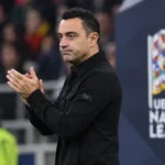 Een coach in donkere jas applaudisseert langs de zijlijn bij een Nations League-wedstrijd.