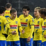 Een groep spelers van KVC Westerlo in gele shirts overlegt op het veld tijdens een avondwedstrijd.