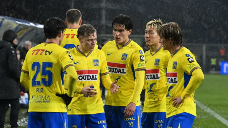 Een groep spelers van KVC Westerlo in gele shirts overlegt op het veld tijdens een avondwedstrijd.