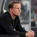 Een coach in zwarte kleding zit geconcentreerd op de bank langs het veld.