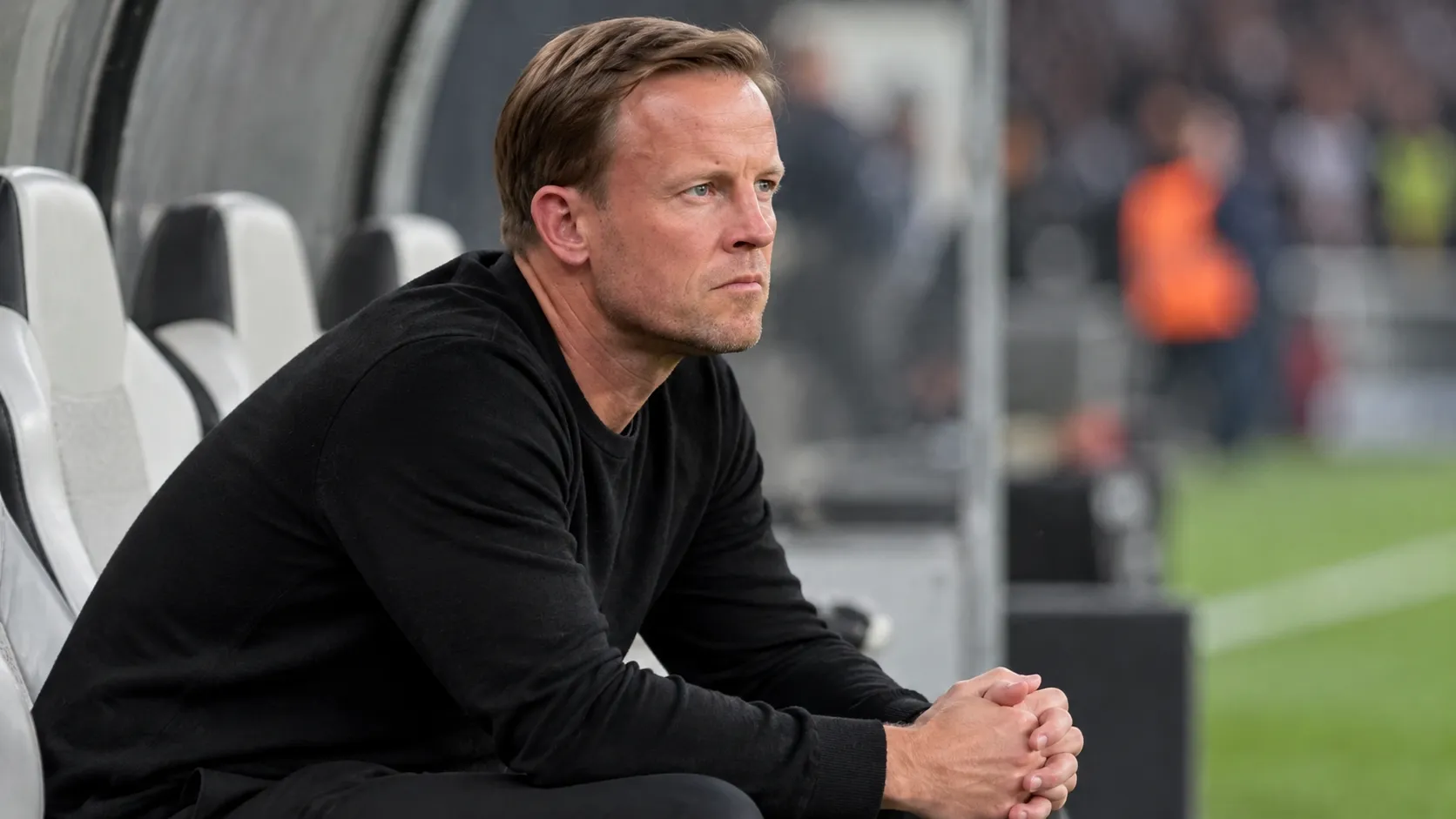 Een coach in zwarte kleding zit geconcentreerd op de bank langs het veld.