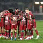 Een groep spelers vormt een hechte huddle op een nat voetbalveld.