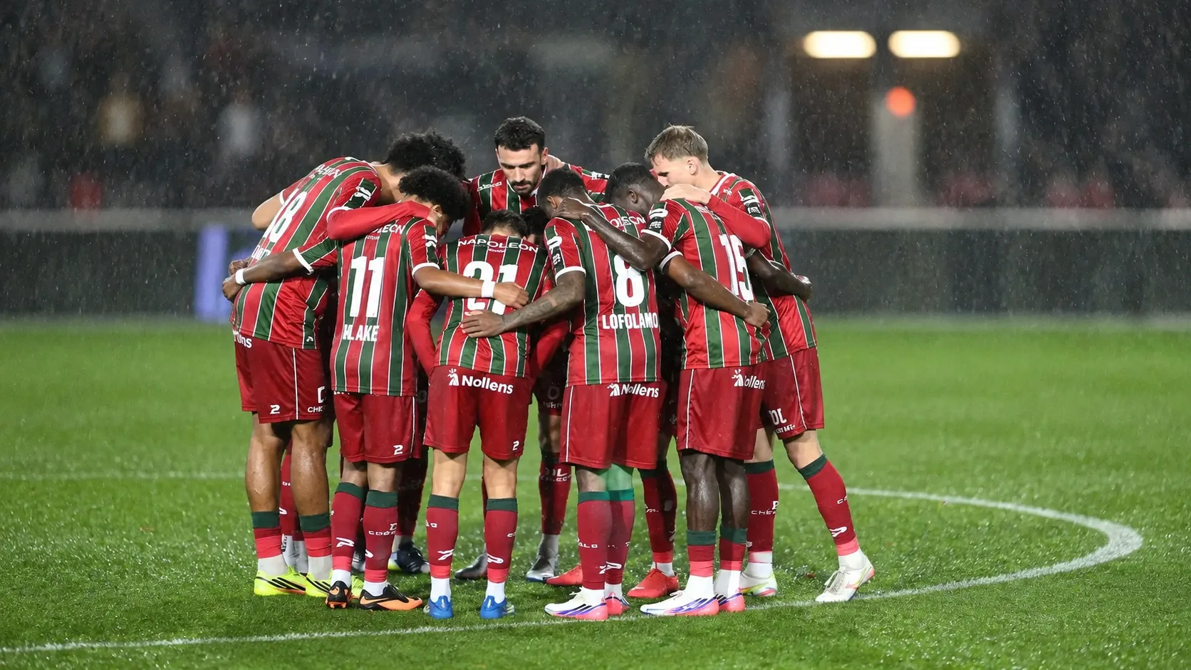 Een groep spelers vormt een hechte huddle op een nat voetbalveld.