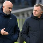 Twee clubmedewerkers in donkere jassen wandelen langs de rand van een voetbalveld.