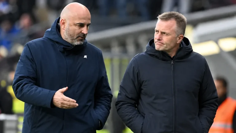 Twee clubmedewerkers in donkere jassen wandelen langs de rand van een voetbalveld.