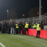 Een groep supporters in donkere jassen bij stewards langs de zijlijn.