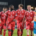 Een groep spelers van KV Kortrijk in rode wedstrijdkleding loopt samen over het veld onder stadionlichten.