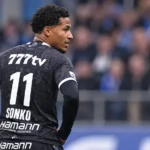 Momodou Sonko in een zwart wedstrijdshirt langs de zijlijn van een voetbalveld.
