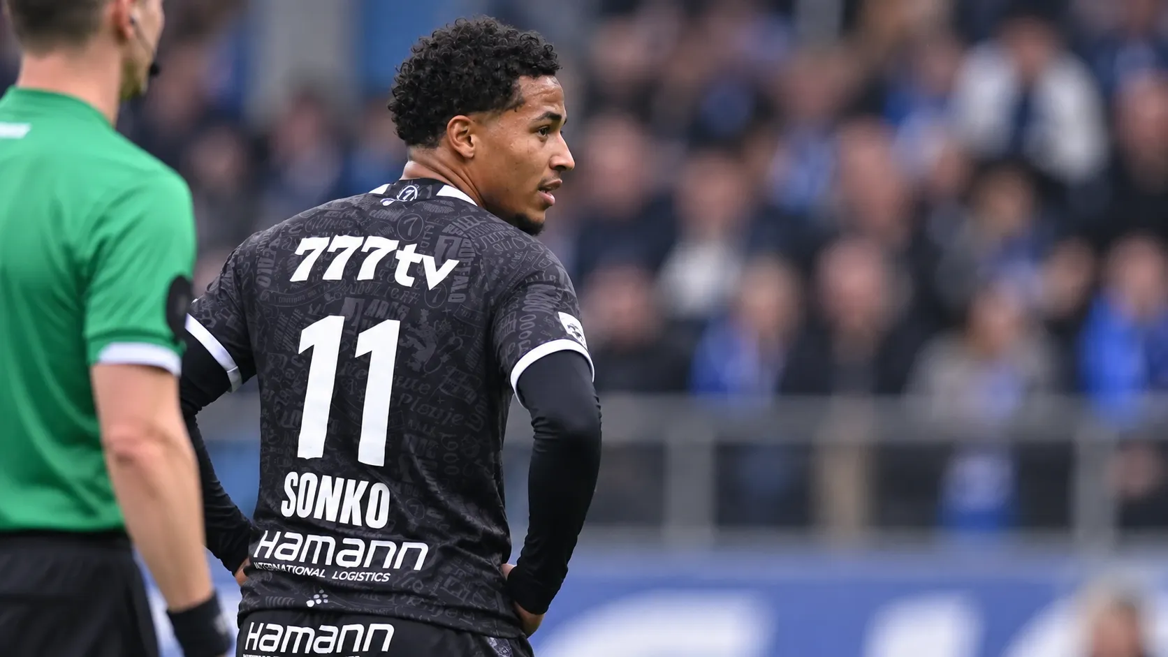 Momodou Sonko in een zwart wedstrijdshirt langs de zijlijn van een voetbalveld.