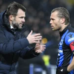 Een trainer in een donkere jas spreekt langs het veld met een speler van Club Brugge.