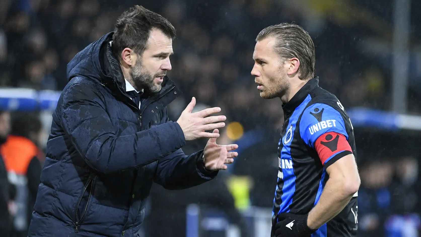 Een trainer in een donkere jas spreekt langs het veld met een speler van Club Brugge.