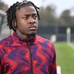 Johan Bakayoko staat geconcentreerd naast een voetbalveld in een trainingsjas.