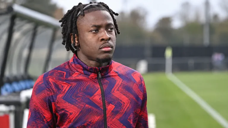 Johan Bakayoko staat geconcentreerd naast een voetbalveld in een trainingsjas.