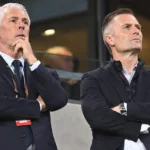 Twee clubofficials in donkere pakken volgen aandachtig de actie vanuit de tribune.