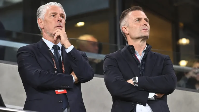 Twee clubofficials in donkere pakken volgen aandachtig de actie vanuit de tribune.
