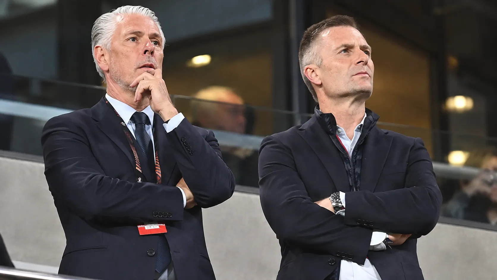Twee clubofficials in donkere pakken volgen aandachtig de actie vanuit de tribune.
