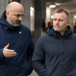 Twee clubofficials in donkere jassen wandelen naast elkaar in een stadionomgeving.