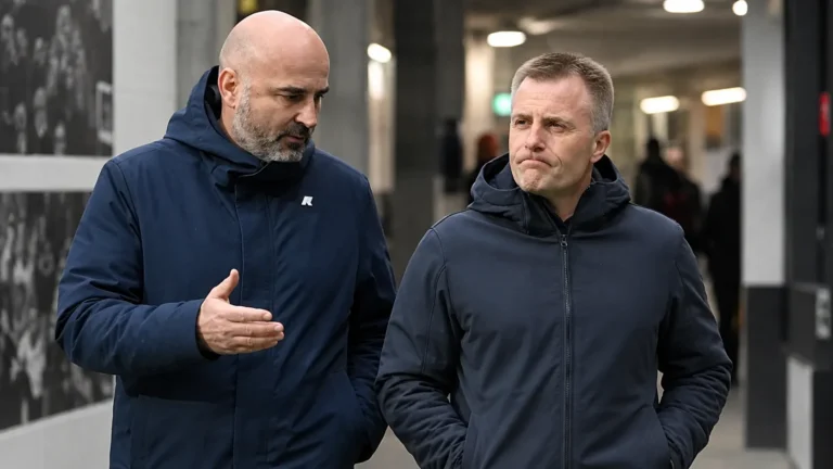 Twee clubofficials in donkere jassen wandelen naast elkaar in een stadionomgeving.