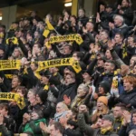 Een volle groep supporters met geel-zwarte sjaals op de tribune.