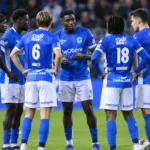 Een groep KRC Genk-spelers in blauwe wedstrijdtenues tijdens een compacte ploegbespreking op het veld.