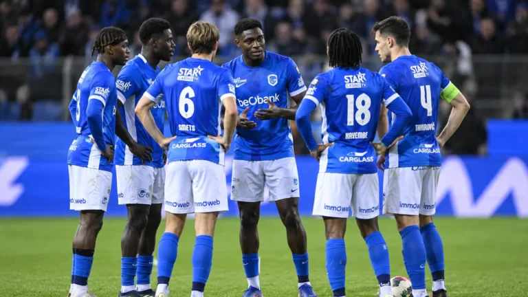 Een groep KRC Genk-spelers in blauwe wedstrijdtenues tijdens een compacte ploegbespreking op het veld.