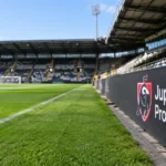 Een leeg stadionvak met een Jupiler Pro League-wedstrijdbord langs het veld.