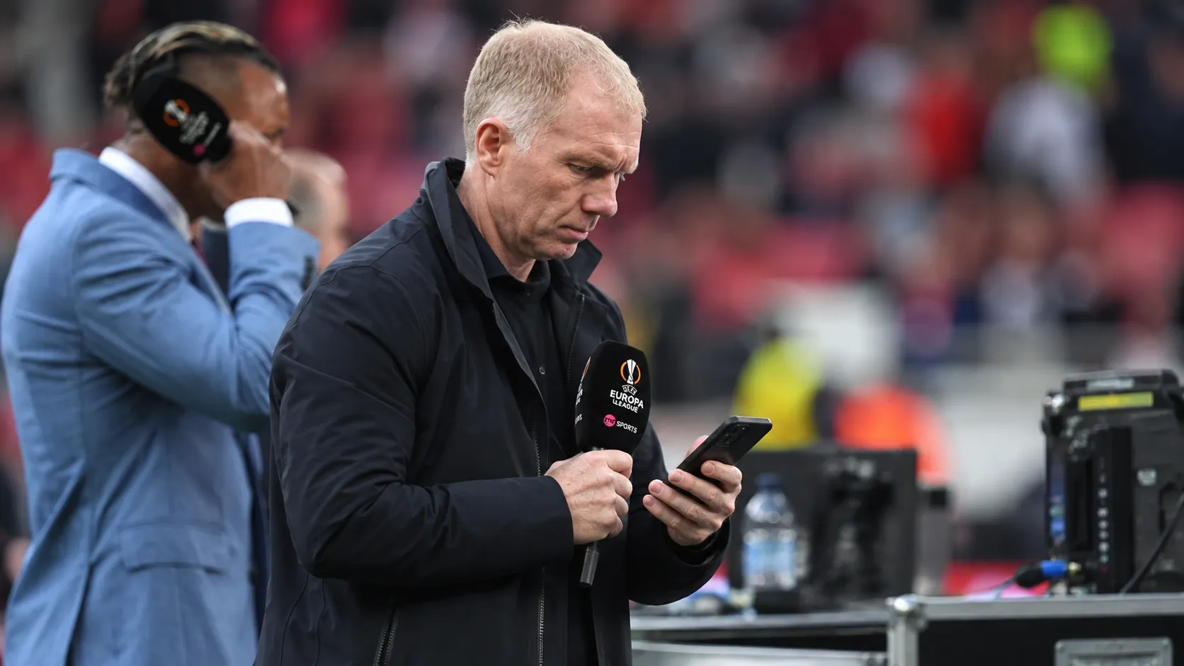 Paul Scholes kijkt op een smartphone aan een mediawerkplek in een voetbalstadion.