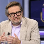 Marc Coucke spreekt in een televisiestudio met een subtiel RSC Anderlecht-embleem op een scherm achter hem.