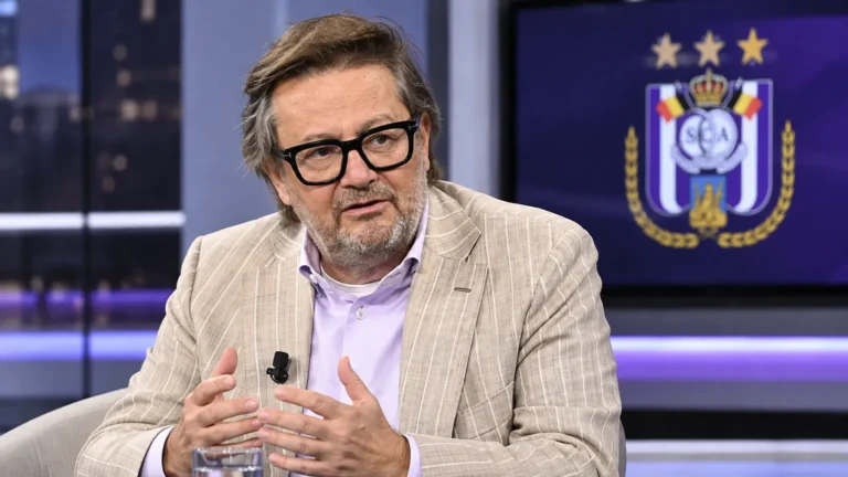 Marc Coucke spreekt in een televisiestudio met een subtiel RSC Anderlecht-embleem op een scherm achter hem.