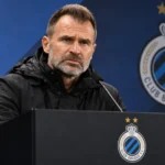 Een Club Brugge-coach in donkere jas aan een lessenaar tijdens een persconferentie.