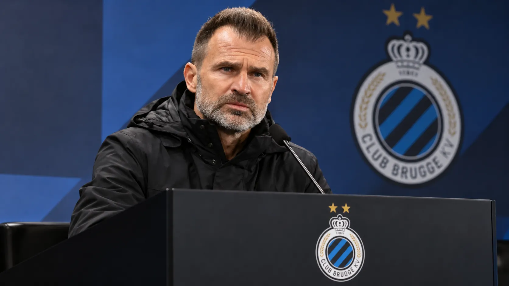 Een Club Brugge-coach in donkere jas aan een lessenaar tijdens een persconferentie.