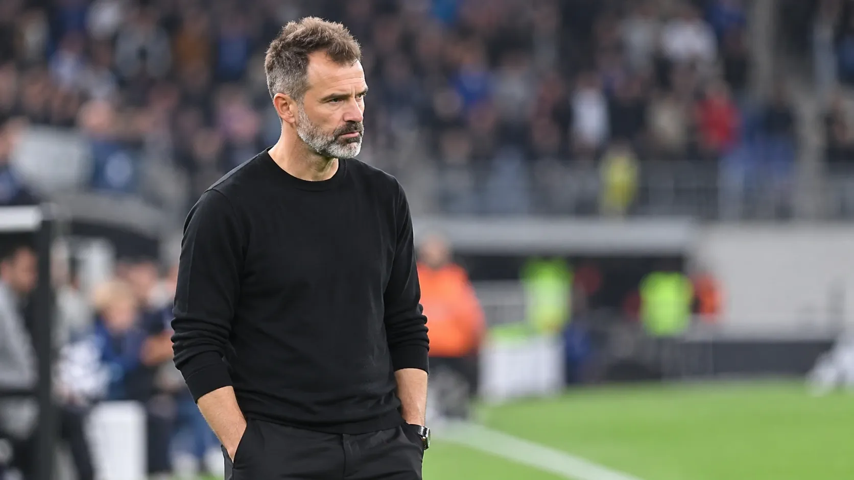Een trainer in een zwarte trui bestudeert het veld vanaf de stadionrand.
